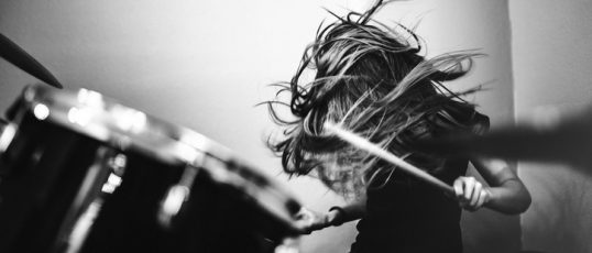 A 9 year old girl has fun playing a drum set in her room, working on songs for her band.  High contrast black and white image.